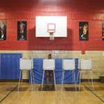A person votes in a gym with a basketball hoop behind them