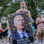 A protester holds a large image of President Biden looking bewildered and surrounded by question mark and thinking face emojis. Under the image are the words 