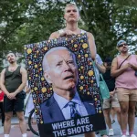 A protester holds a large image of President Biden looking bewildered and surrounded by question mark and thinking face emojis. Under the image are the words 