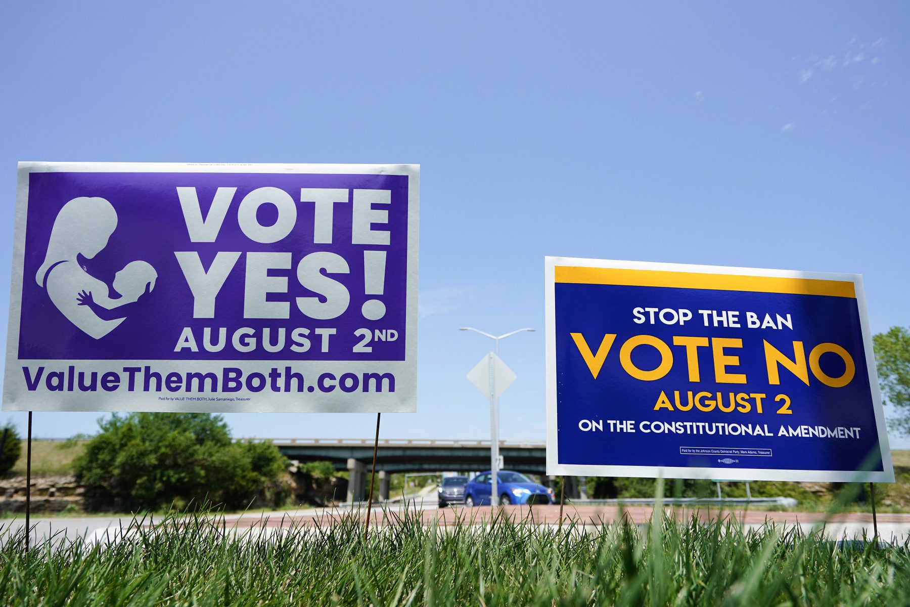 Two signs next to each other. A purple one reads "vote yes! August 2nd" with a silhouette of a person holding a baby. The other reads "stop the ban! Vote no August 2nd."