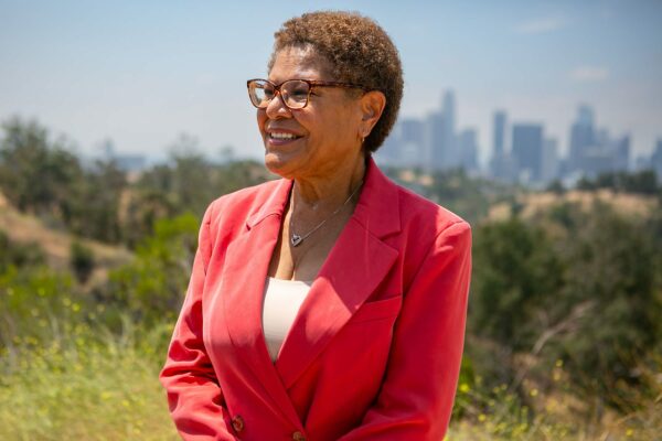 Karen Bass looks out and smiles. A view of downtown Los Angeles can be seen behind her.