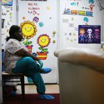 A woman puts on protective gloves and slippers before starting work at her daycare center inside her home. The walls are decorated with Dr. Seuss quotes and characters and informative posters.