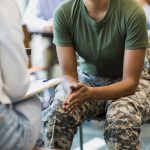 A close up photo of an unrecognizable mid adult female soldier as she puts her hands together and leans forward in her seat.