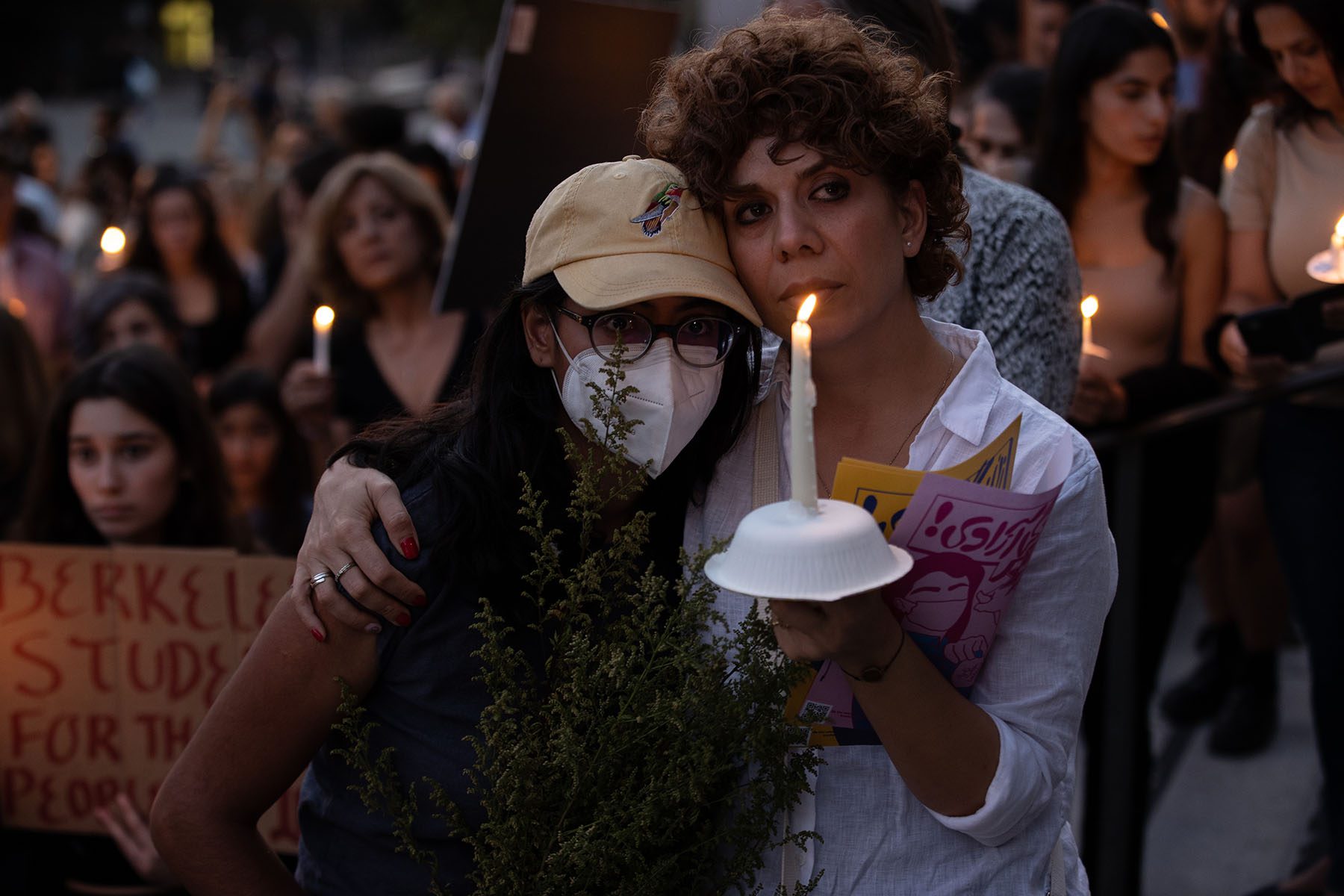 A young woman holds a bouquet and hugs an older woman holding a candle at a candlelit vigil. Behind them, a large crowd can be seen.
