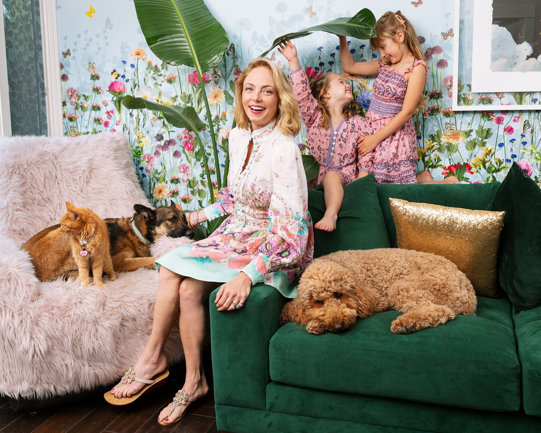 Louisette Geiss poses in her living room. On her left an orange cat and a dog lay on a fluffly couch. Behind her, her two daughters play with a plant. On her right, another dog takes a nap on a velvet couch.