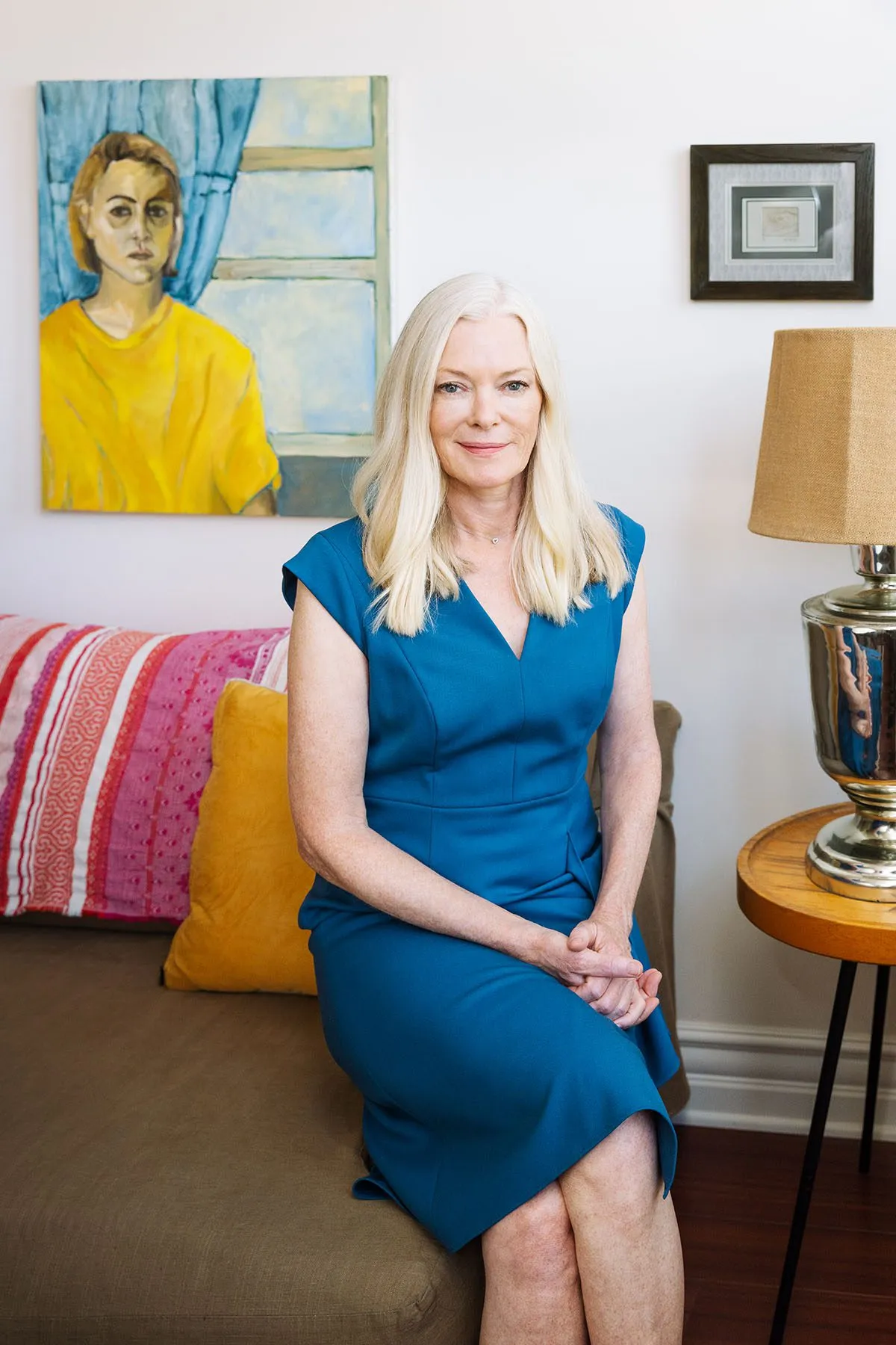 Caitlin Dulany sits cross legged on the edge of a couch. Behind her is a portrait of a women by a window.