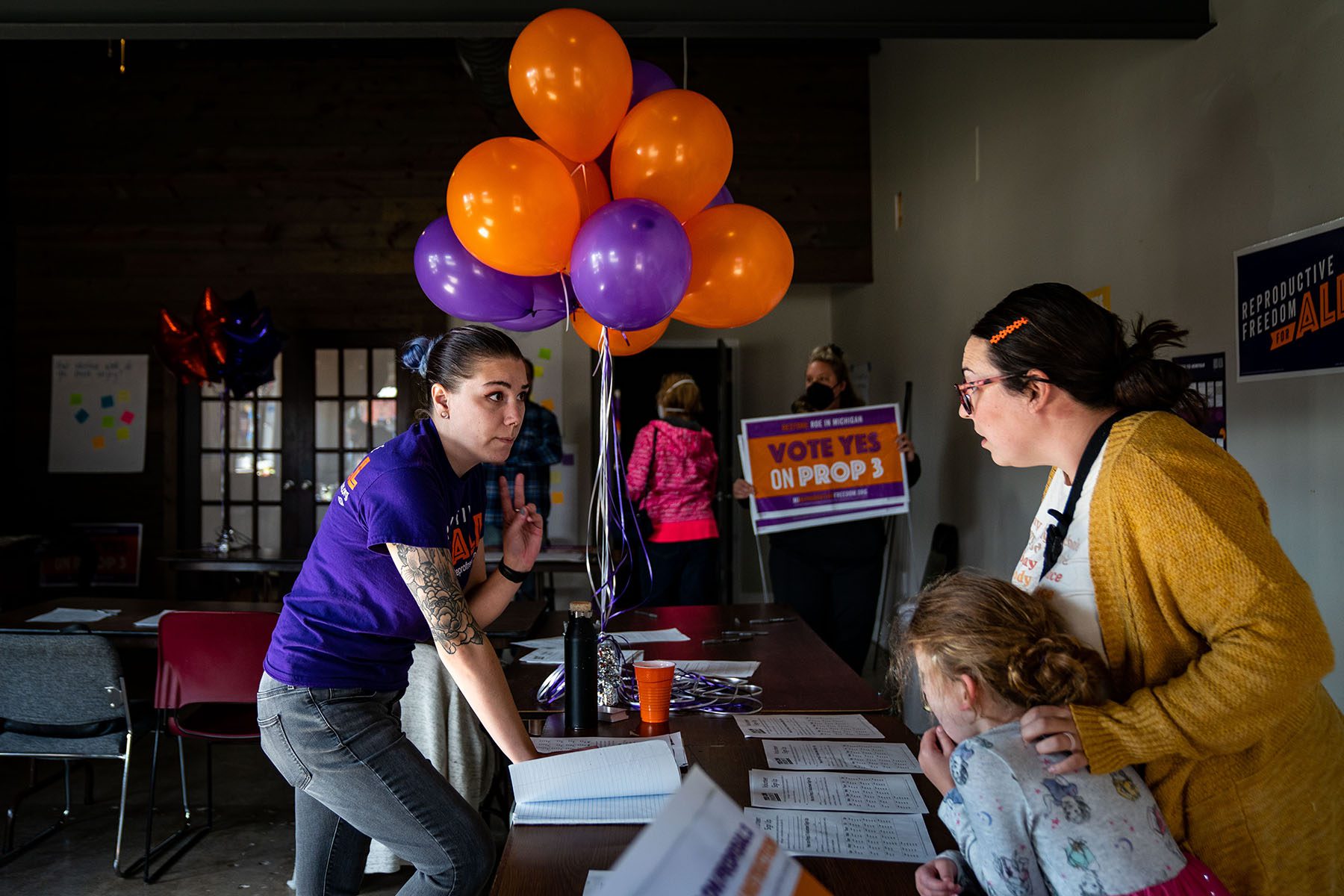 An organizer helps a volunteer register and pick up signs at a Reproductive Freedom for All campaign office.  Purple and orange balloons decorate a table and "Agree Yes to Pillar 3" Farm signs can be seen in the .  Background.