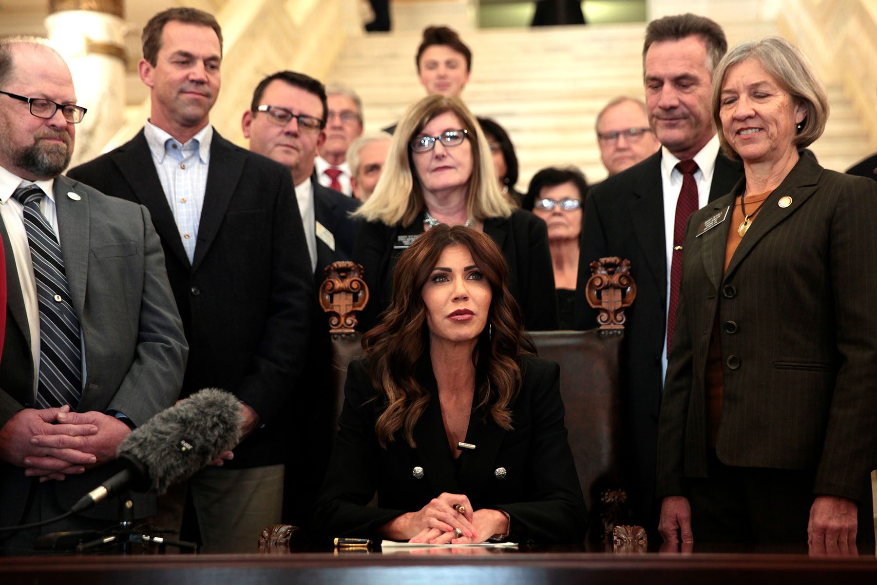 Kristi Noem sits at a desk surrounded by lawmakers as she signs an anti-trans bill into law.