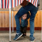 A child peaks at the photographer from under his mother's legs as he waits for his mother to cast her ballot.