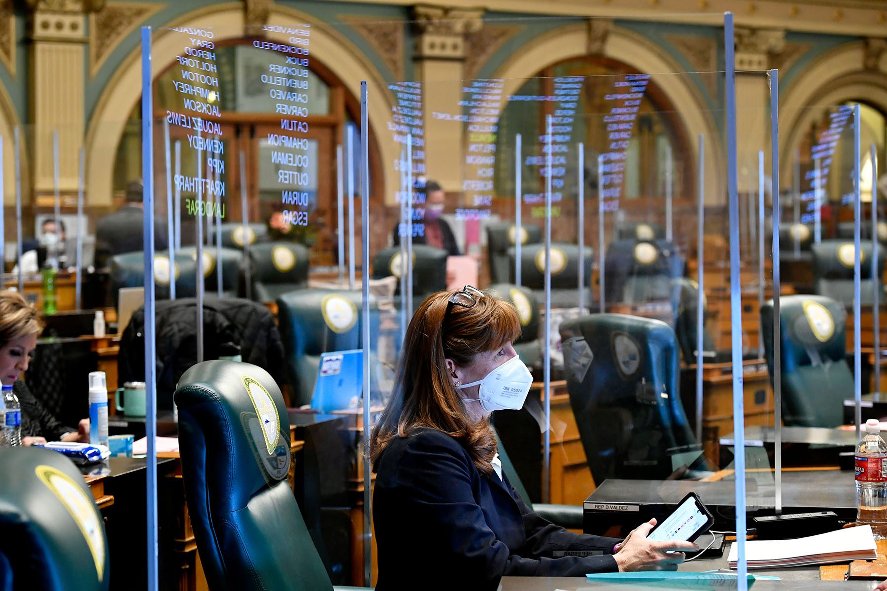 Rep. Julie McCluskie wears a face mask and is surrounded by protective plastic barriers reflecting the names of Colorado representatives from the monitor on the wall in the House chambers at the Colorado State Capitol during an emergency legislative session.
