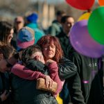 Tearful people embrace outside a vigil at All Souls Unitarian Church. Rainbow colored balloons can be seen in the foreground.