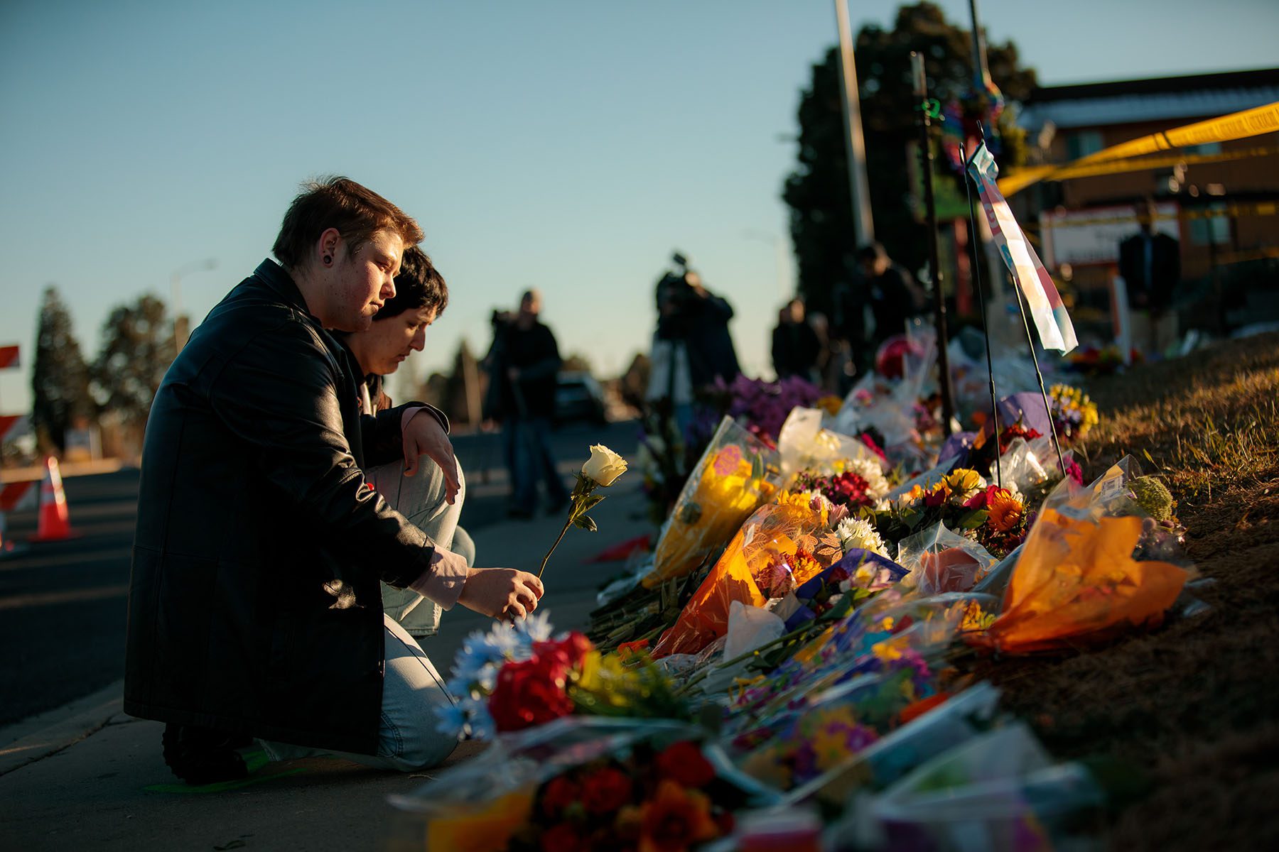 People leave flowers at a growing memorial. Flags and bouquets of flowers are scattered on the ground, police tape still visible in the background.