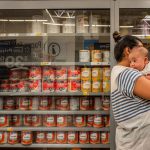A mother holds her baby in front of a locked shelving unit stocked with baby formula.