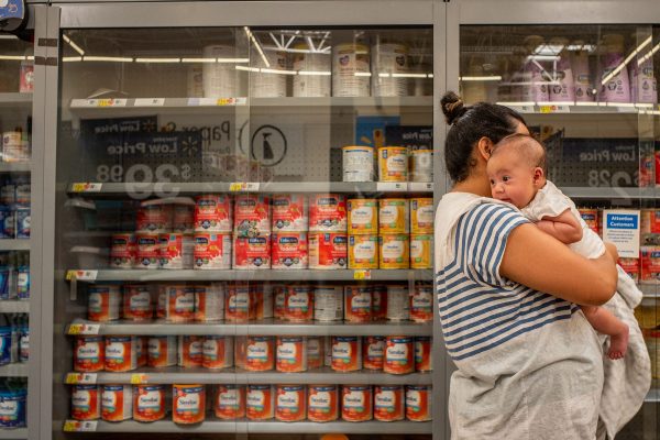 A mother holds her baby in front of a locked shelving unit stocked with baby formula.