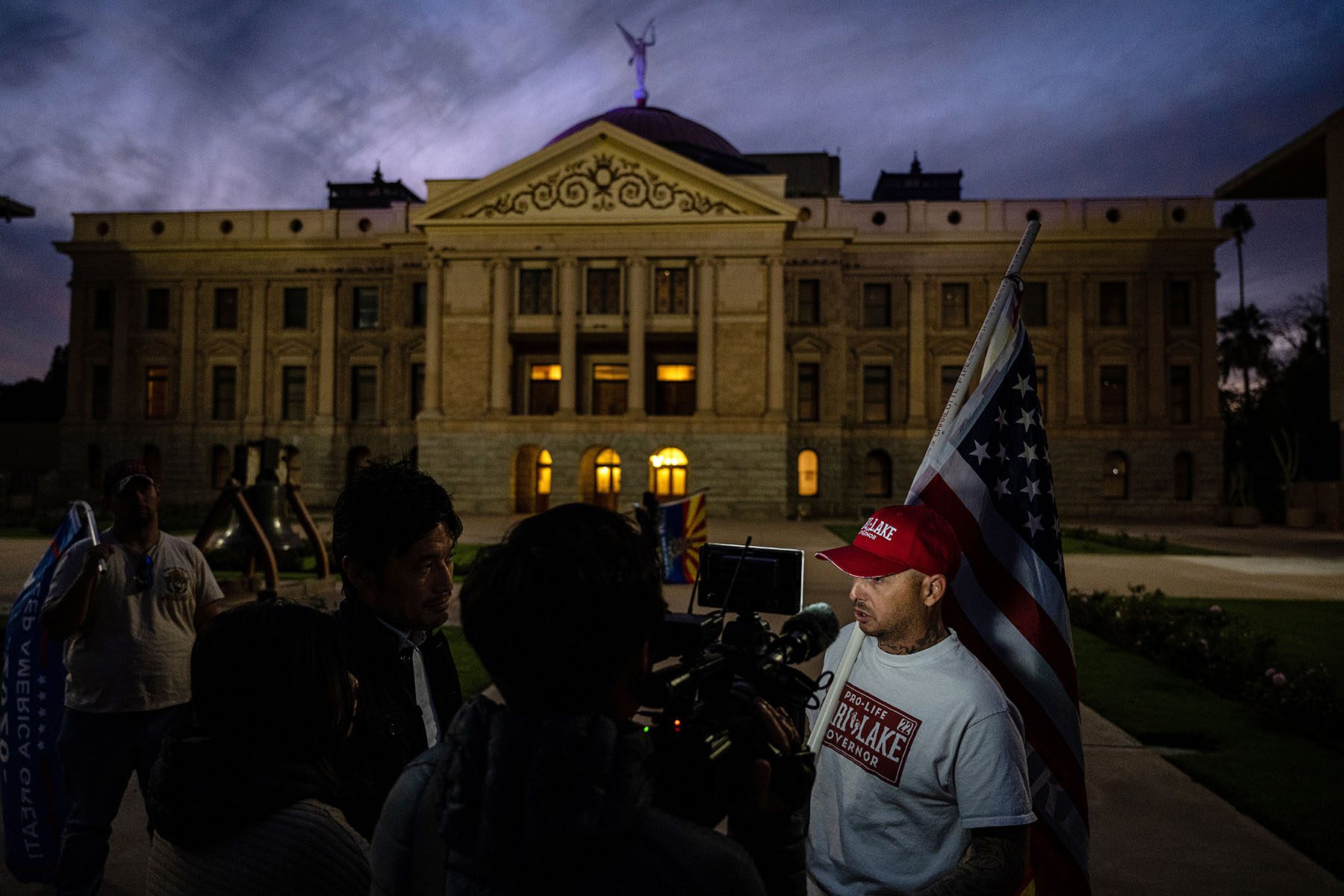 A Kari Lake supporter holding an American flag speaks to a news crew in front of the Arizona State Capitol building.