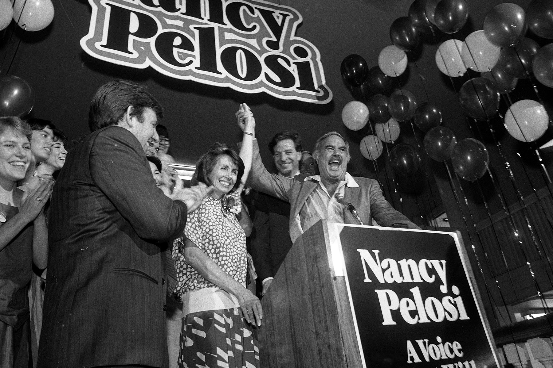 Nancy Pelosi raises her fist from a podium. Supporters clap and cheer around her.