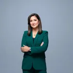 Headshot of Amna Nawaz crossing her arms and smiling in a green suit over a grey background..