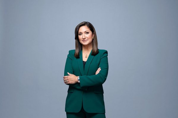 Headshot of Amna Nawaz crossing her arms and smiling in a green suit over a grey background..