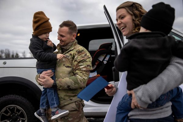 A U.S. Army Staff Sargent holds his son in his arms and smiles as his spouse holds their other child.