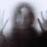 Back lit image of the distorted silhouette of a person with their hands pressed against a glass window.