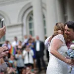 Two women embrace after getting married as people around them clap, cheer and take pictures.