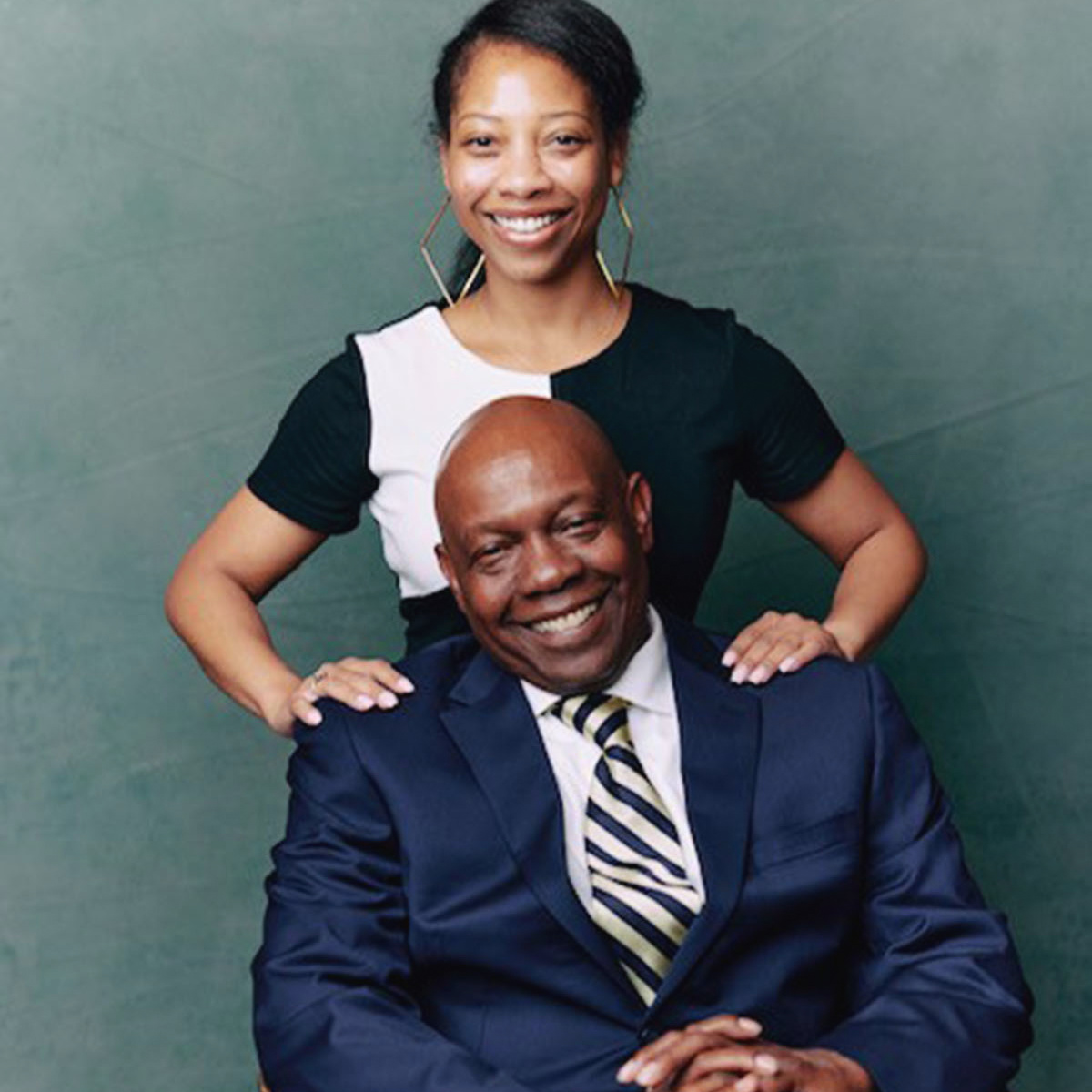 A portrait of Malia Fredrick and her father: he is sitting on a chair and smiling, she is behind him with her hands on his shoulders.