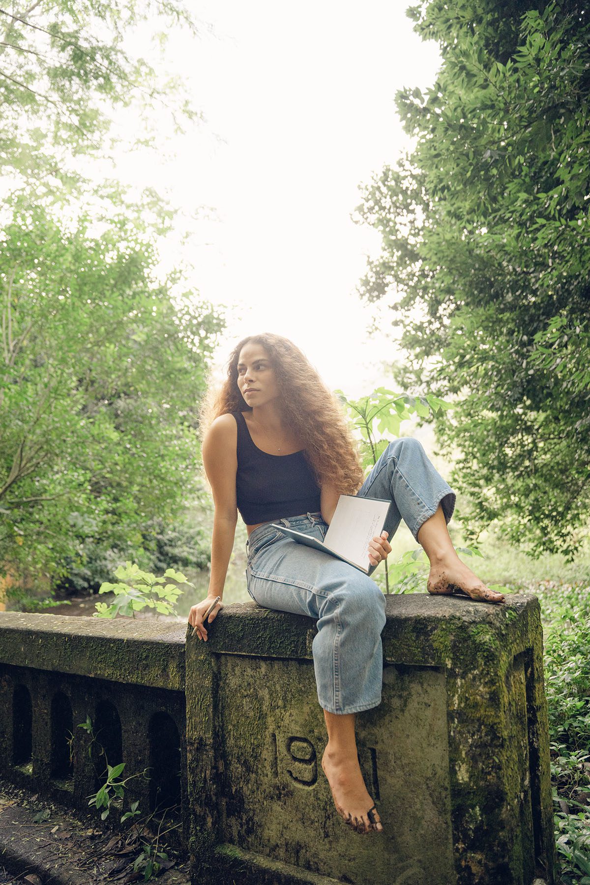 Sage Lenier poses for a portrait on a bridge. She is barefoot and holding an open notebook with one hand.