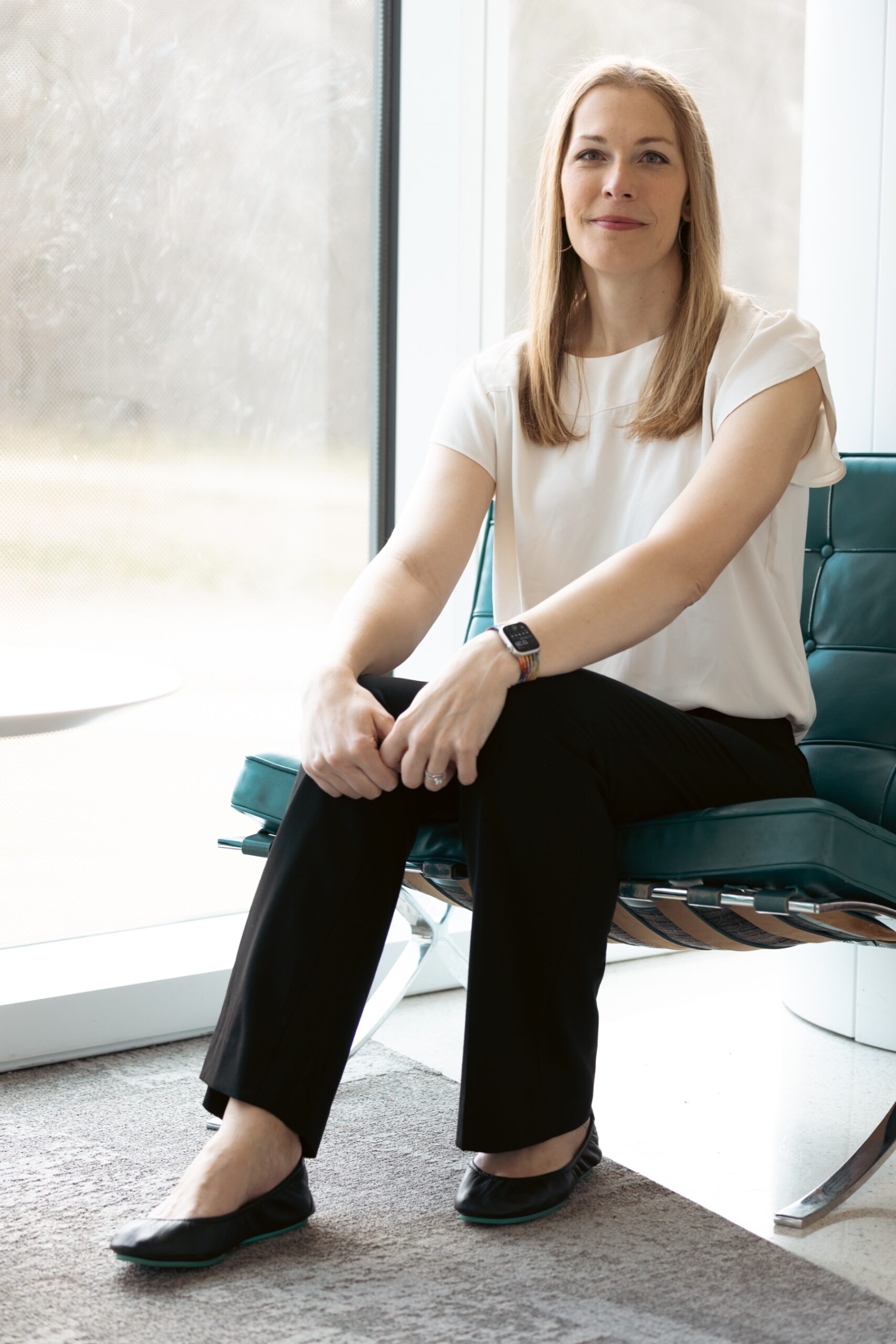 A woman with medium-blonde hair wears a white blouse and black slacks while sitting in a sunny office space.