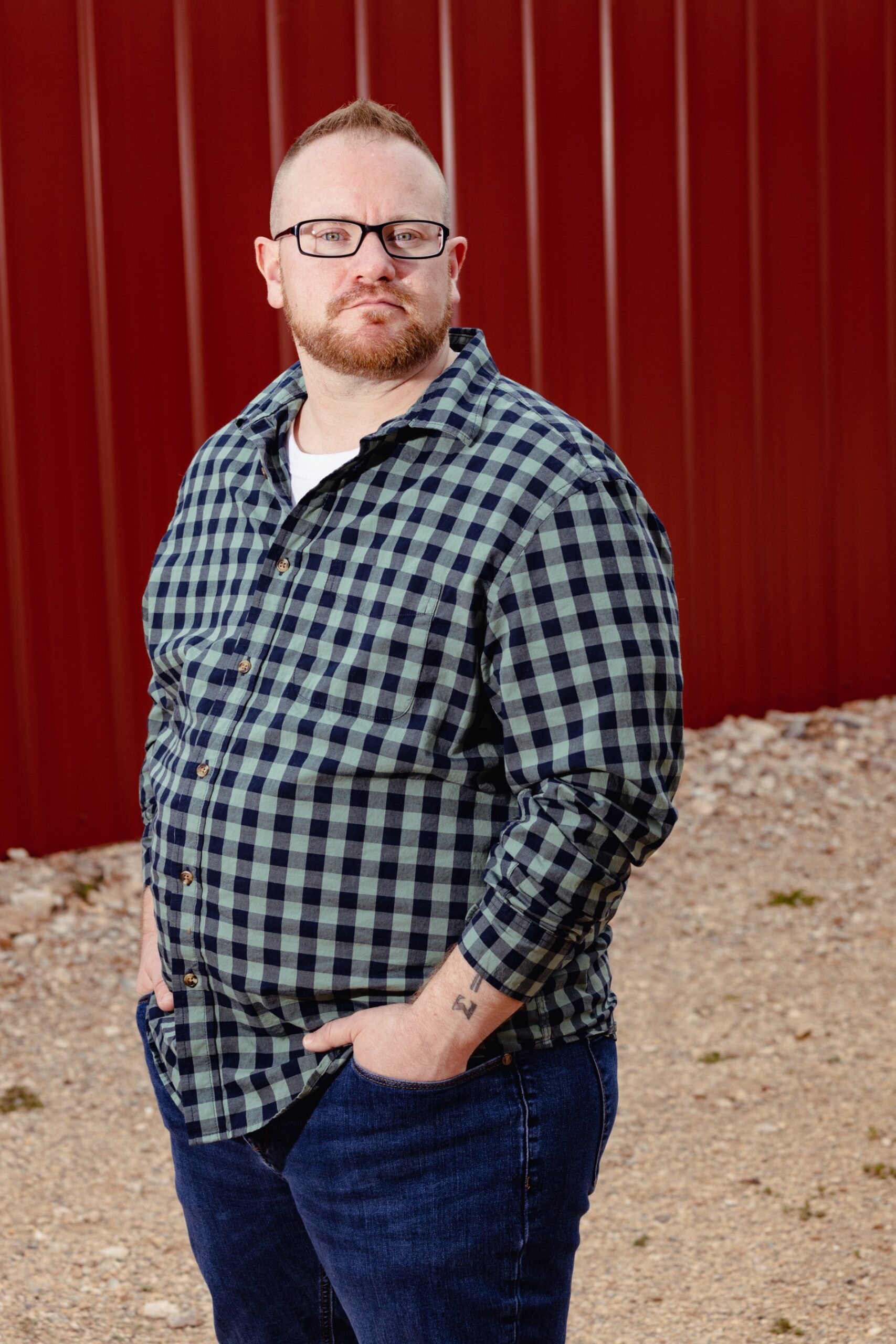 A man with reddish hair and a beard wears a green checked shirt, jeans, and rectangular glasses. He's standing in a dirt area in front of a red wall.
