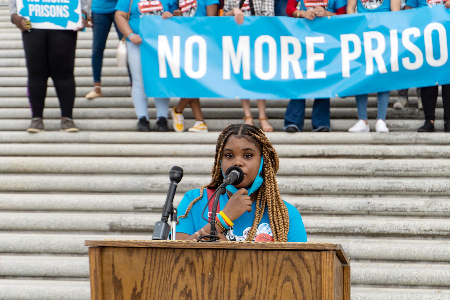 Tamia Cenance speaks at a podium as activists hold a banner that reads "no more prisons!" behind her.