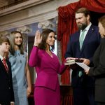Senator Katie Britt, surrounded by her family is sworn in by Vice President Kamala Harris. Katie Britt raises her hand and smiles as her other hand is placed on a bible held by her husband. Behind her, her son and daughter are smiling.