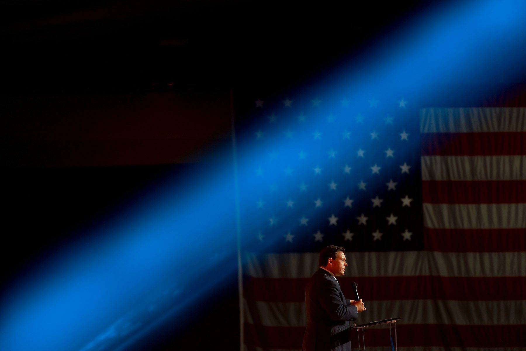 Florida Gov. Ron DeSantis speaks at a podium. A bright blue stage light passes above his head. Behind him is a large American flag.
