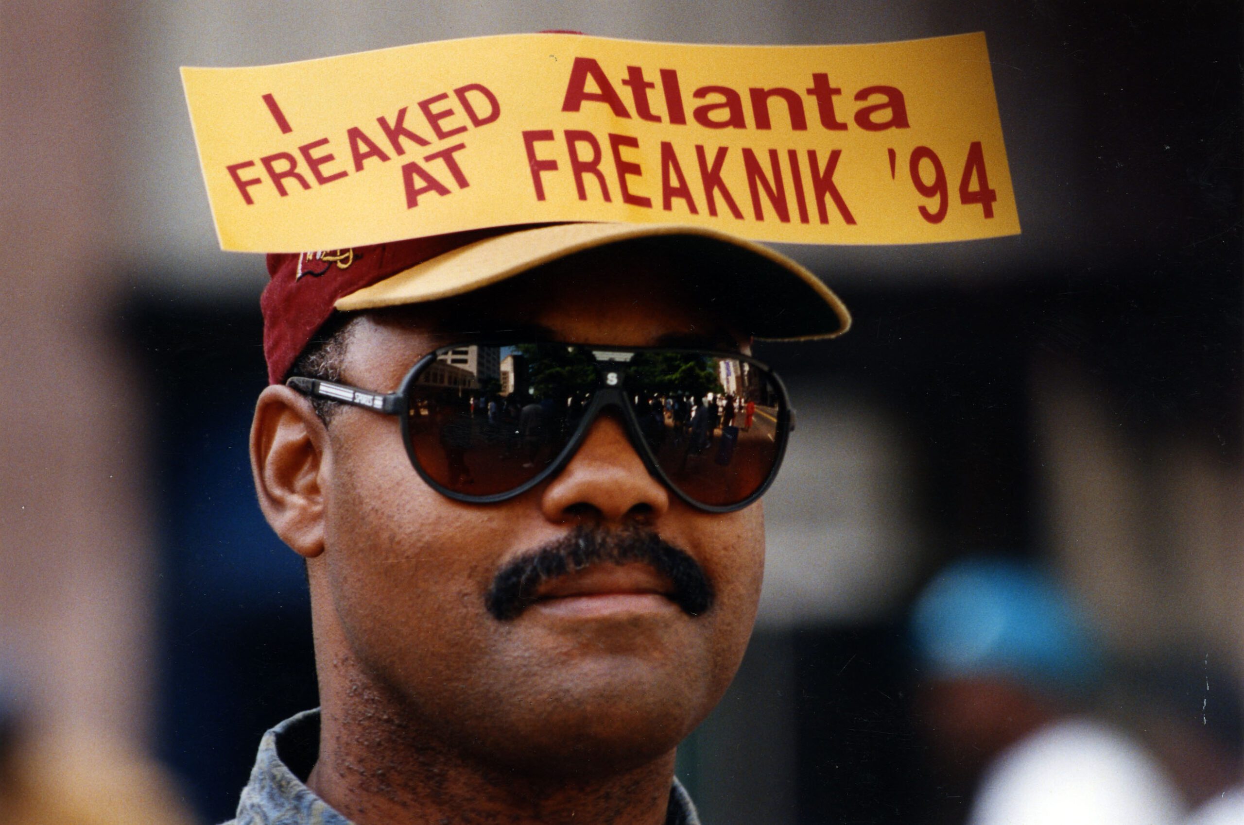Man wearing sunglasses reflecting the city off of them and wearing a sign on his hat that reads, "I Freaked at Atlanta Freaknik '94"