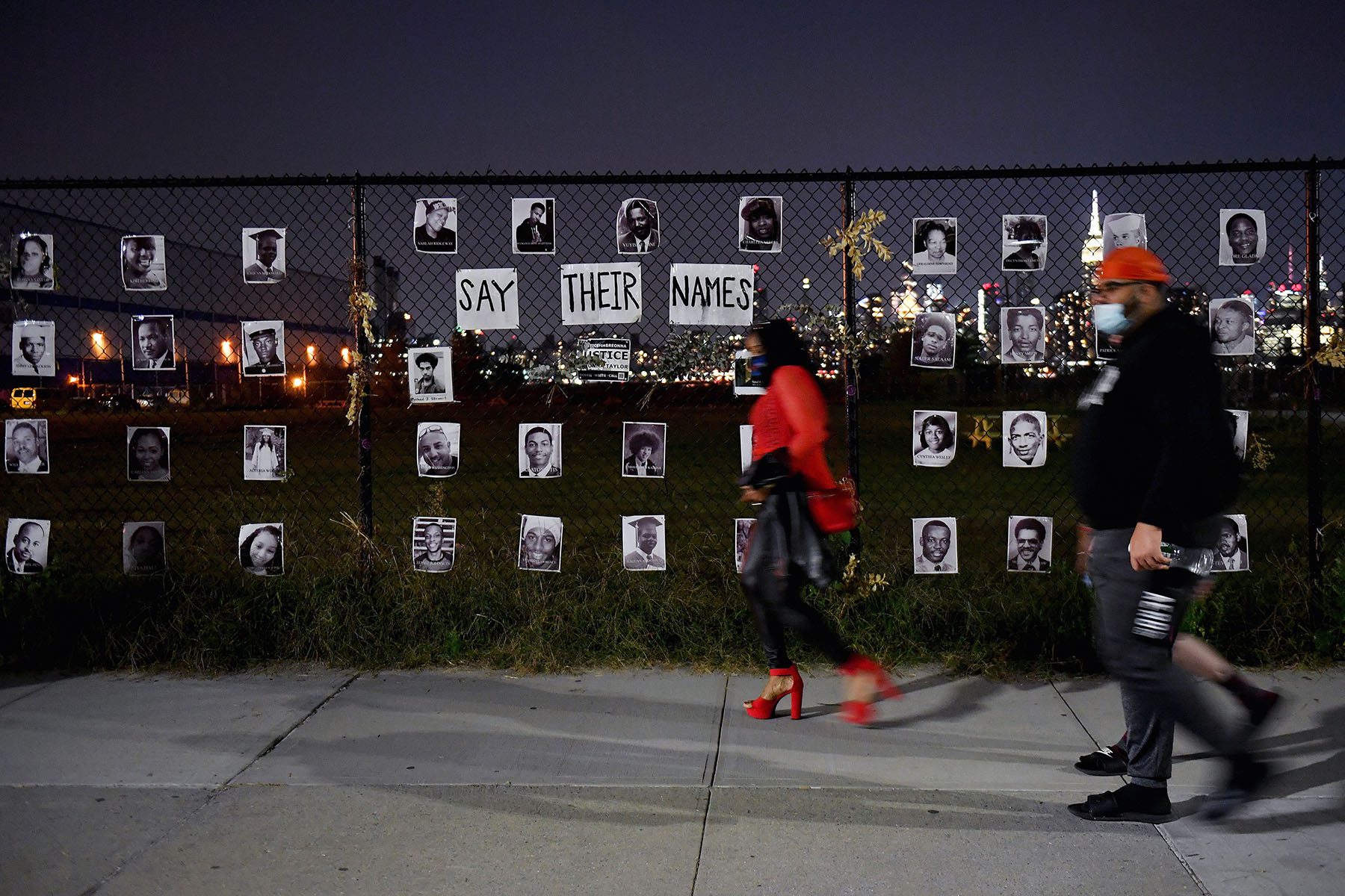 people walk by a fence at night time that has the photos of people killed by the police posted on it.