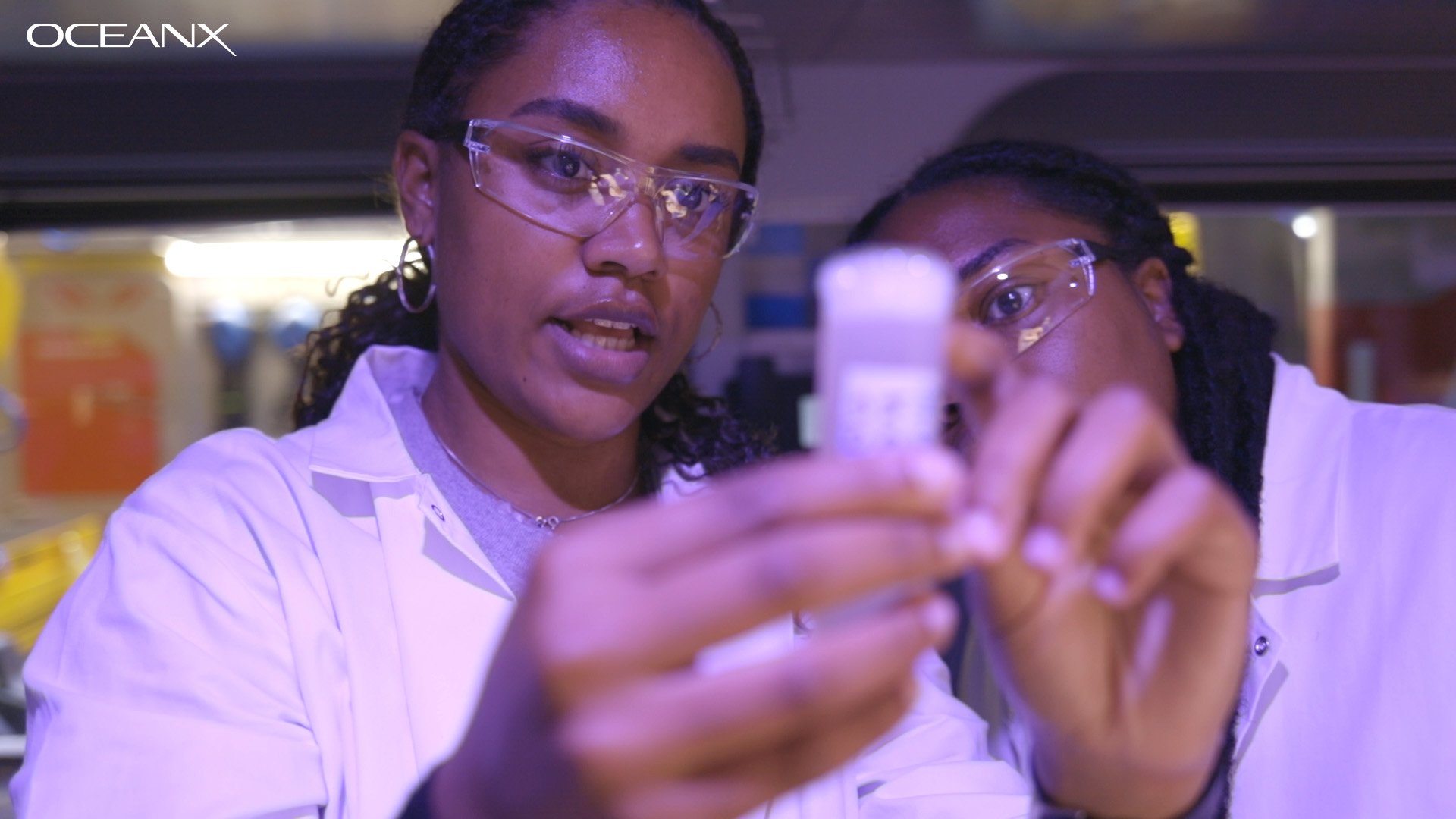 Two black women wearing goggles and lab coats speak to each other in a lab aboard OceanXplorer.
