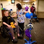 A caregiver checks a resident in a wheelchair's blood pressure in a nursing home hallway.