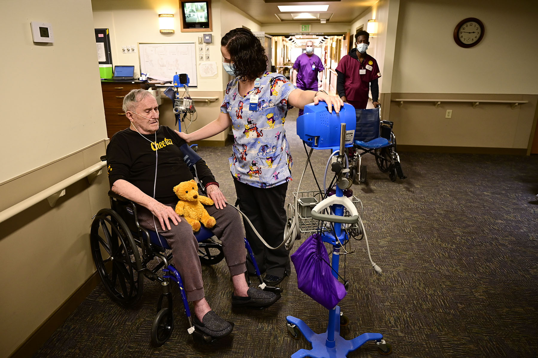 A caregiver checks a resident in a wheelchair's blood pressure in a nursing home hallway.