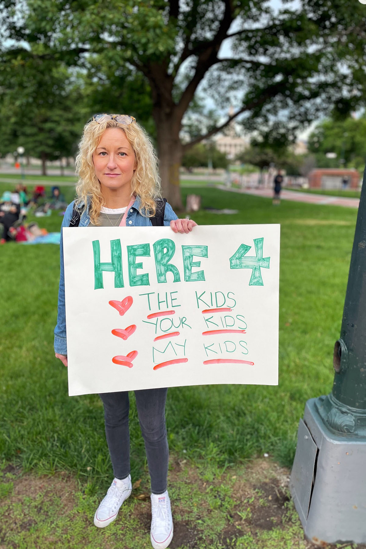 Katie MacKenzie, a participants in the sit in poses for a portrait near the Colorado State Capitol. She holds a sign that reads "Here 4 The Kids, Your Kids, My Kids."