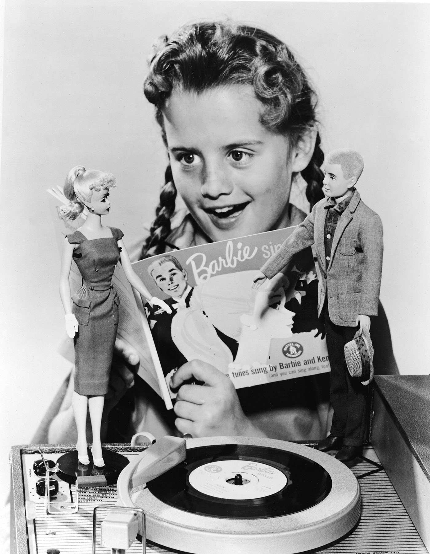 A girl in pigtails sings along with a 7" record called 'Barbie Sings' which plays on a portable phonograph player in 1961. Two dolls, Barbie and Ken, stand on the phonograph.