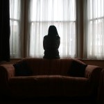 A silhouetted woman looks out a window in a dark room.
