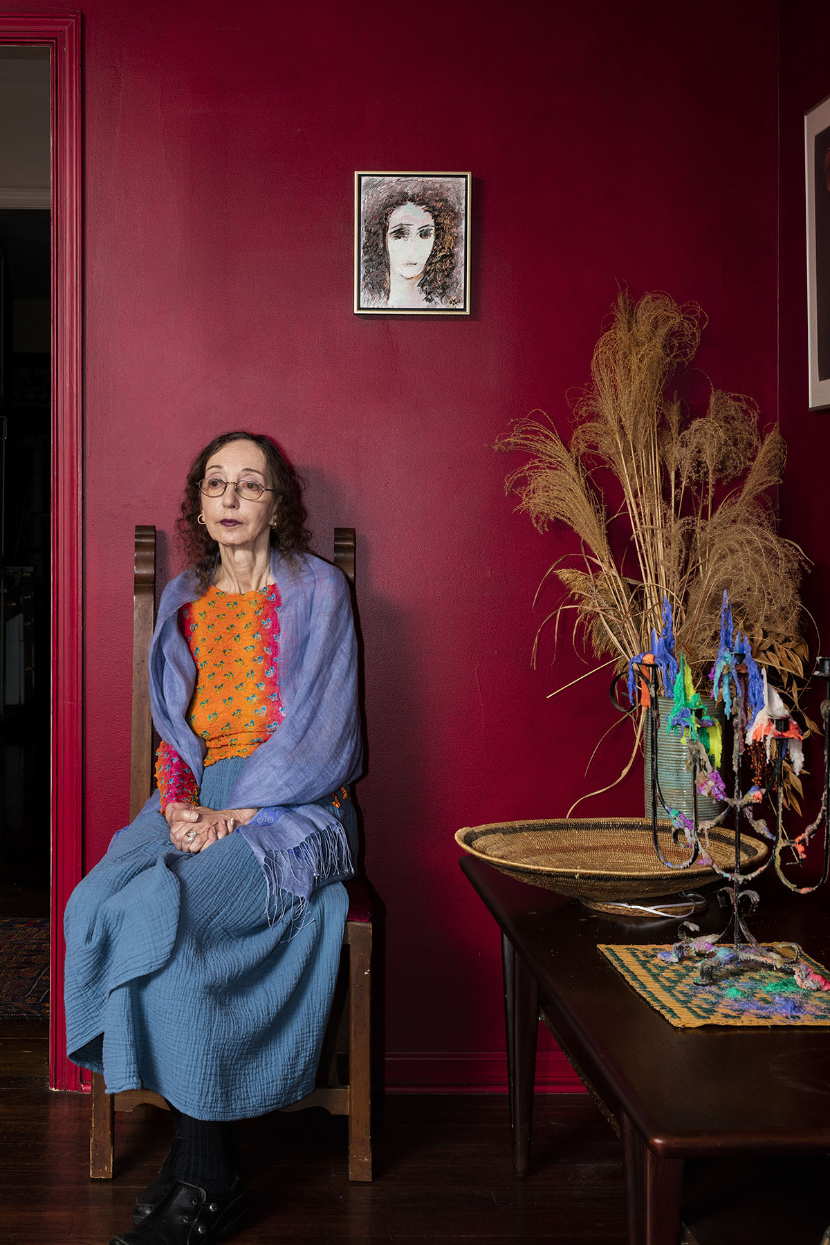 Portrait of Joyce Carol Oates sitting on a chair in front of a red wall.