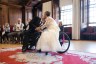 A bride and groom, who are wheelchair users have their first kiss after exchanging their vow while the wedding guests clap and celebrate them in the background.