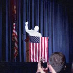 Photo collage of a silhouetted figured raising their fist while standing at a podium as people clap in the foreground. The figure at the podium is surrounded by American flags.