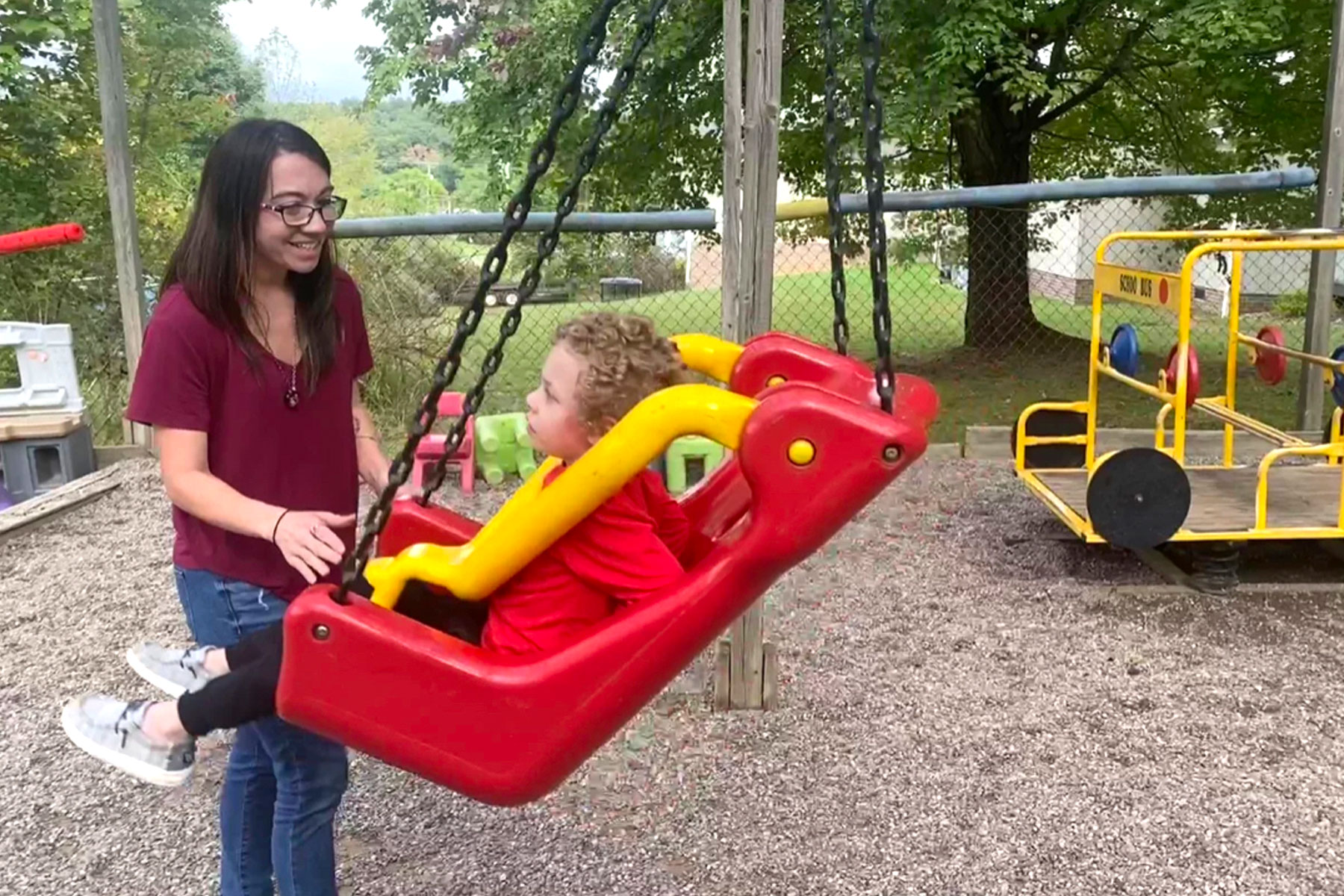 A women stands in front of a child swinging in a red toddler swing on a playground.