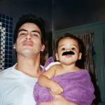 A dad and his child share a moment after a bath. The baby is wearing a fake mustache.