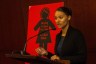 T Ortiz stands at a podium speaking into a microphone. Behind her is a red poster that reads, 