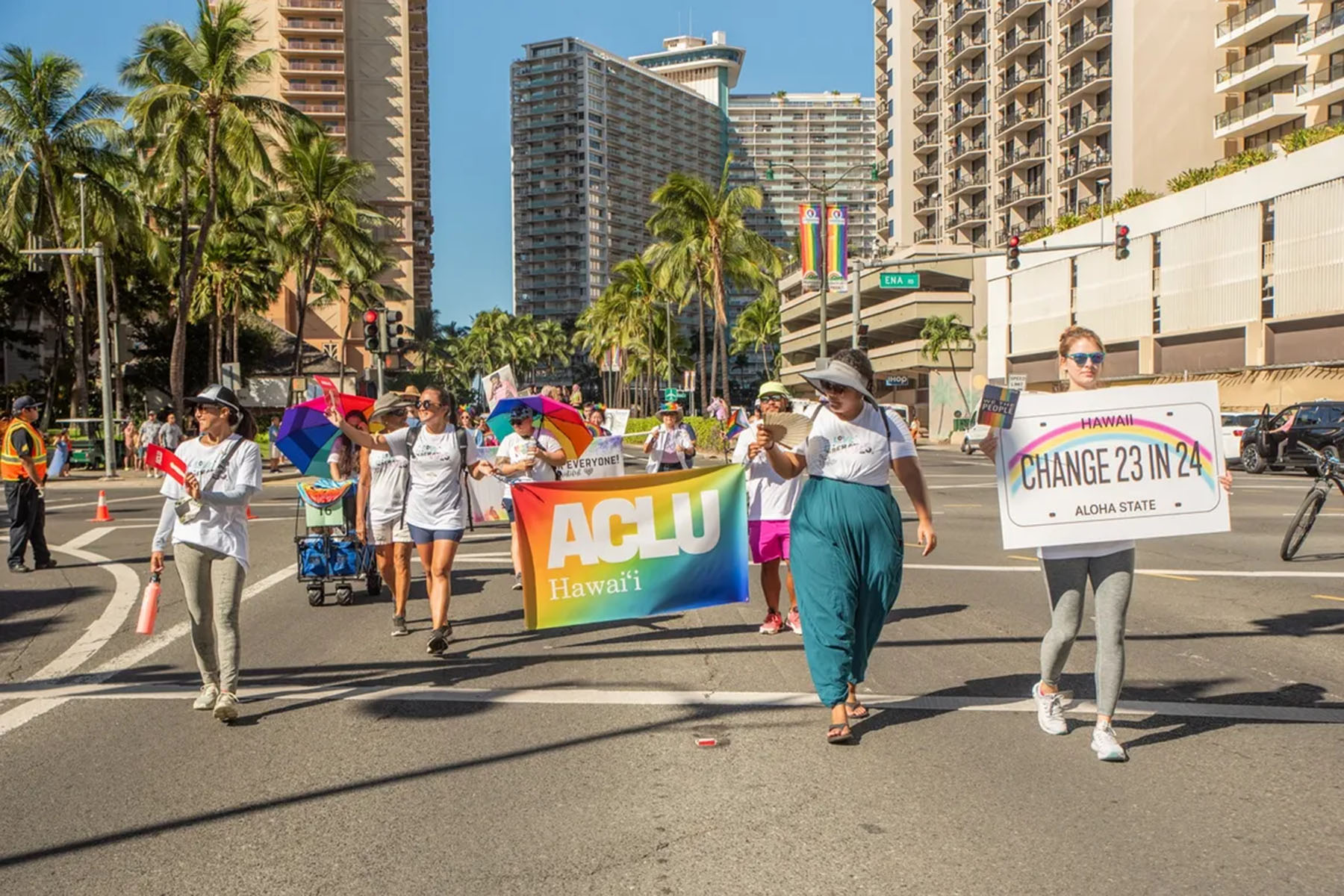 People walk down the streets holding signs for ACLU Hawaii and one that reads "Change 23 in 24" during Hawaii Pride.