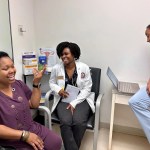 A smiling woman gestures at two medical professionals who are also smiling.