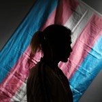 A silhouetted person stands in front of a trans pride flag.
