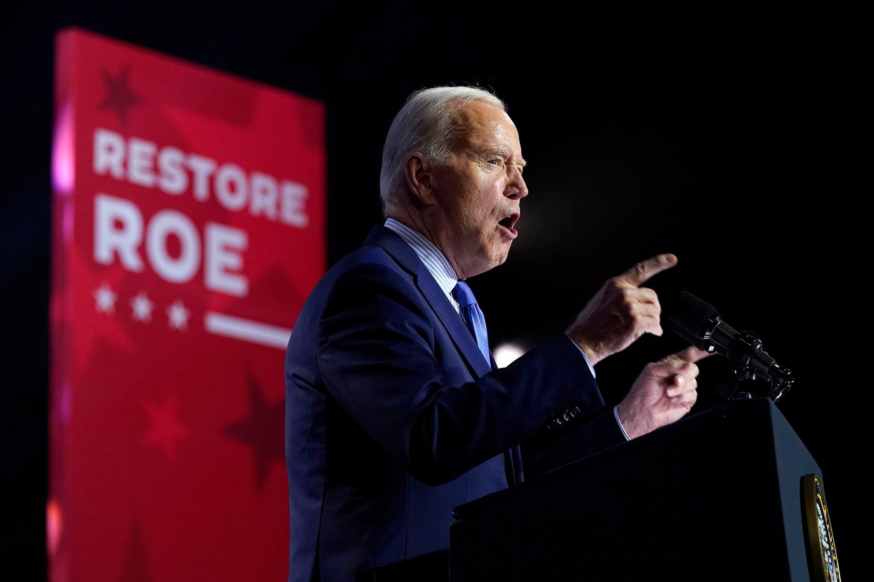 President Biden speaks during an event. Behind him a panel reads "Restore Roe."